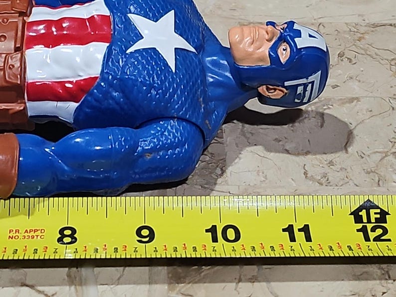 May include: A blue and red Captain America action figure with a white star on his chest. The figure is lying on its side and is approximately 8 inches long. A yellow measuring tape is in the foreground with the numbers 8, 9, 10, 11, and 12 visible.