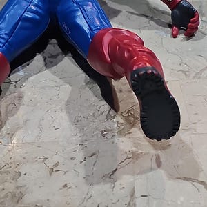 May include: A red, white, and blue action figure of a superhero in a crawling pose. The figure has a black boot with a textured sole.