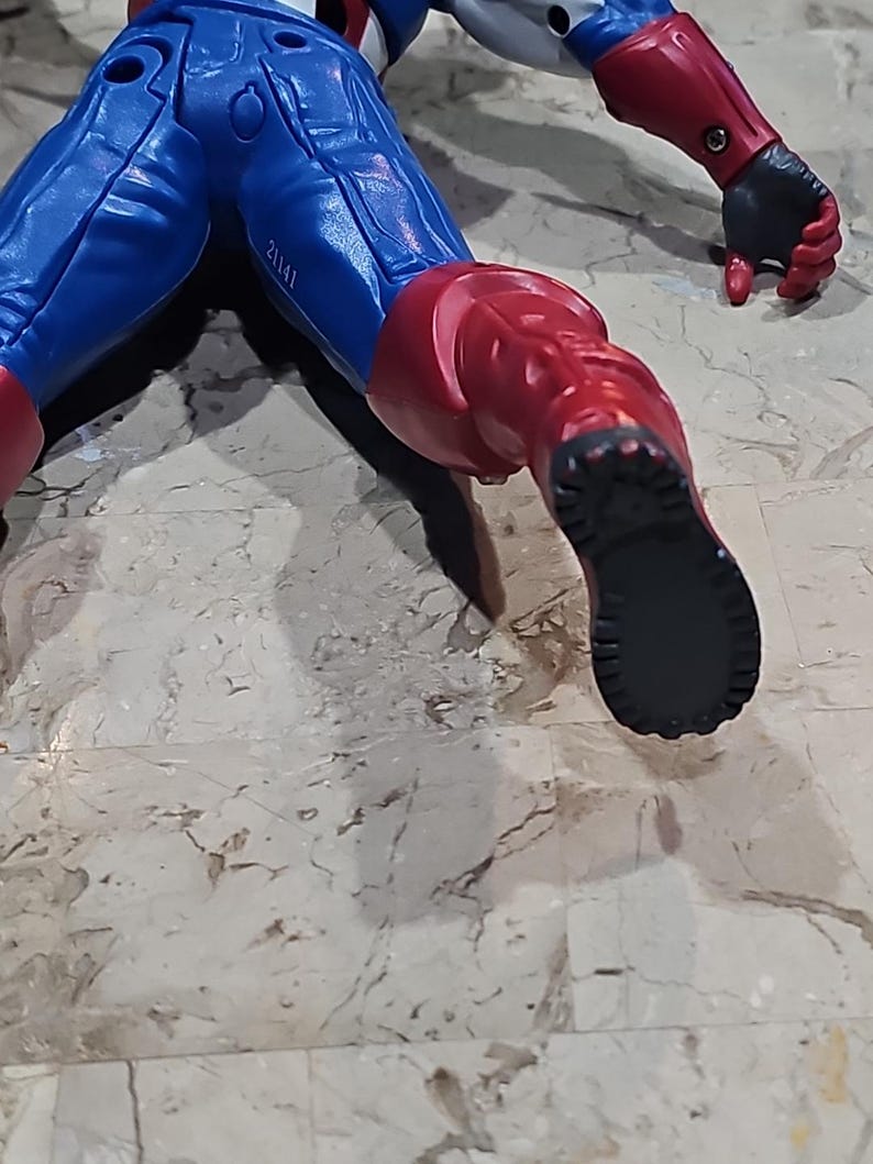 May include: A red, white, and blue action figure of a superhero in a crawling pose. The figure has a black boot with a textured sole.