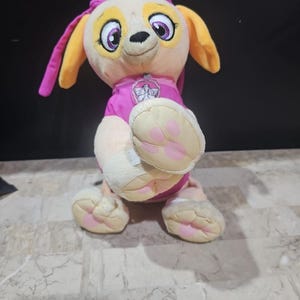 Op de afbeelding: Een pluche speelgoed van Skye van Paw Patrol. Ze draagt een roze en witte outfit met een pootprint op haar borst. Ze heeft een roze strik op haar hoofd en haar ogen zijn open.