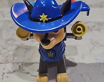 May include: A blue and brown toy dog wearing a blue sheriff's hat with a yellow star. The dog is holding two yellow and black toy guns.