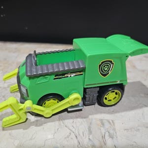 Puede incluir: Camión de juguete verde con un brazo de garra amarillo y un logotipo de huella de pata. El camión tiene una parte trasera extraíble y una garra que funciona.