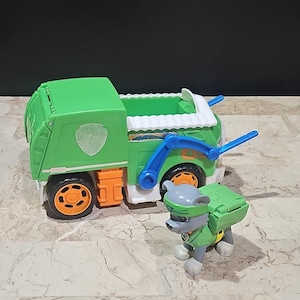 May include: Green toy garbage truck with a white and blue cab and a gray and green toy dog figure. The truck has a paw print logo on the side.