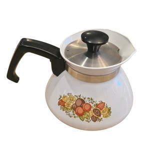 May include: White enamel coffee pot with a black handle and lid. The pot features a floral design with orange, yellow, and brown fruit and flowers. The pot has a stainless steel lid and a gold-colored band.