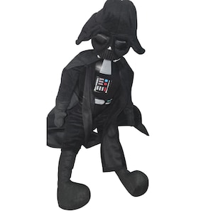 May include: A plush toy of a character wearing a black helmet, a black cape, and a black suit. The chest panel features a rectangular design with red, blue, and white accents. The figure has long, dangling arms and legs with rounded black feet.