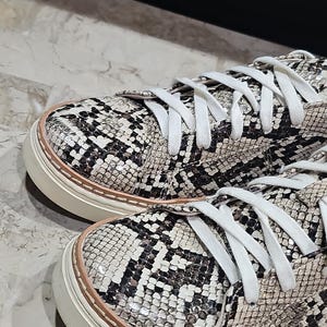 May include: Close-up of a pair of sneakers with a snakeskin pattern in black, white, and tan. The shoes have white laces and a cream-colored sole with a light brown trim. The sneakers are casual footwear.