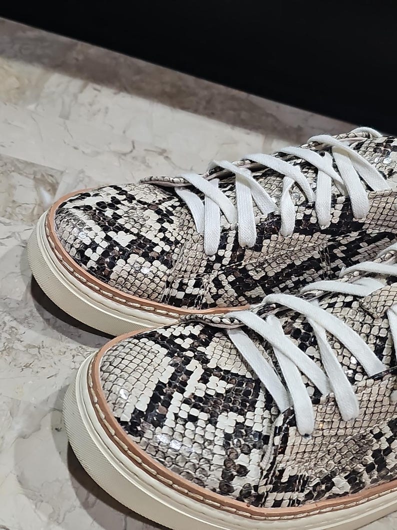 May include: Close-up of a pair of sneakers with a snakeskin pattern in black, white, and tan. The shoes have white laces and a cream-colored sole with a light brown trim. The sneakers are casual footwear.