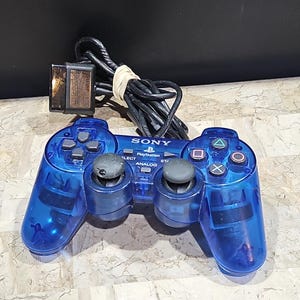 Könnte beinhalten: Ein blauer, durchsichtiger PlayStation-Controller mit einem schwarzen Kabel und einer schwarzen Speicherkarte. Der Controller hat ein PlayStation-Logo und die Wörter "PlayStation" und "Analog" sind darauf gedruckt.