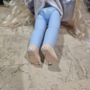 May include: A doll with long blonde hair wearing a blue and white dress and blue pants. The doll is sitting on a light brown surface.