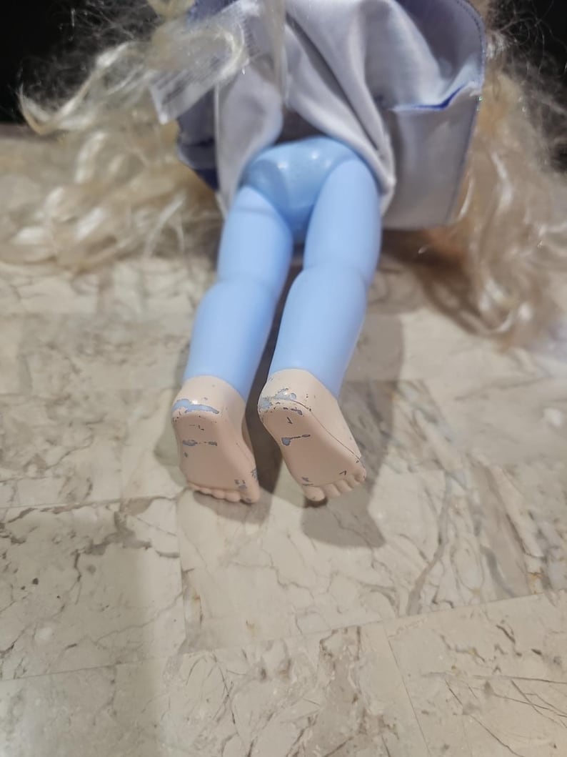 May include: A doll with long blonde hair wearing a blue and white dress and blue pants. The doll is sitting on a light brown surface.
