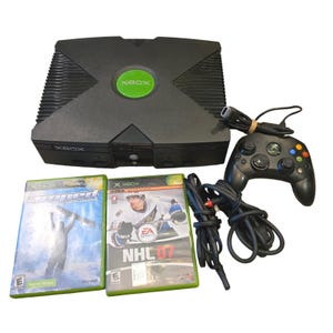 May include: A black original Xbox video game console with a green circular logo, one black wired controller, and two game cases titled Amped and NHL 07. The console features a large X-shaped top design with side vents and front controller ports.