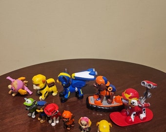 May include: A collection of colorful plastic toy figurines of dogs from the Paw Patrol cartoon series. The figurines are in various poses and include a yellow dog with a blue and white jetpack, a red dog with a red and white snowboard, and a purple dog with a pink and white helicopter.