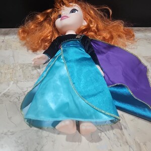 Puede incluir: Una mu&ntilde;eca de pelo rojo con un vestido azul y morado con ribete dorado. La mu&ntilde;eca lleva una capa de terciopelo negro y est&aacute; tumbada sobre una superficie blanca.