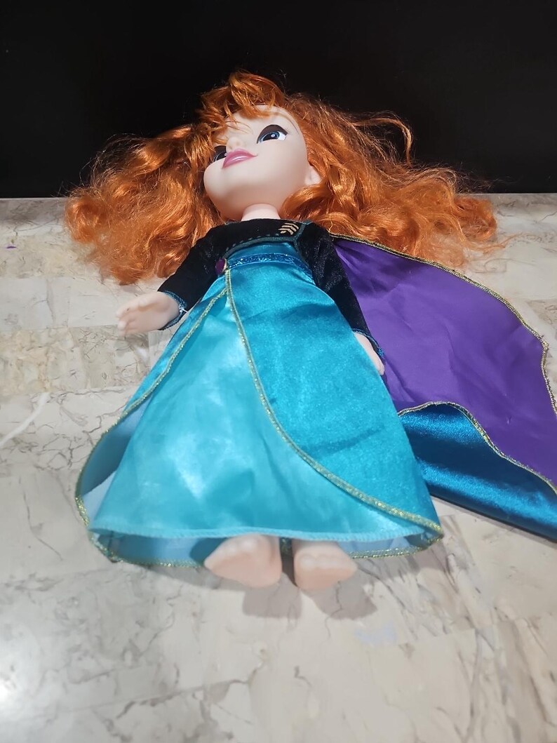 Puede incluir: Una mu&ntilde;eca de pelo rojo con un vestido azul y morado con ribete dorado. La mu&ntilde;eca lleva una capa de terciopelo negro y est&aacute; tumbada sobre una superficie blanca.