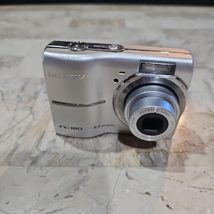 Puede incluir: Una cámara digital Olympus FE-180 plateada con una resolución de 6,0 megapíxeles y un objetivo de zoom óptico 3x. La cámara tiene un cuerpo plateado con un objetivo negro.