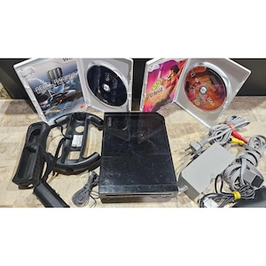 Peut inclure: Une console Nintendo Wii noire avec deux boîtiers de jeu, un accessoire de volant et deux manettes. Les boîtiers de jeu sont ouverts, affichant les disques de jeu et les illustrations de "Rebel Raiders" et "Zumba". Câbles et alimentation inclus.