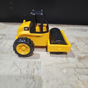 Puede incluir: Veh&iacute;culo de construcci&oacute;n de juguete amarillo con una cabina negra y un rodillo negro. El veh&iacute;culo tiene la palabra "CAT" impresa en el lateral.