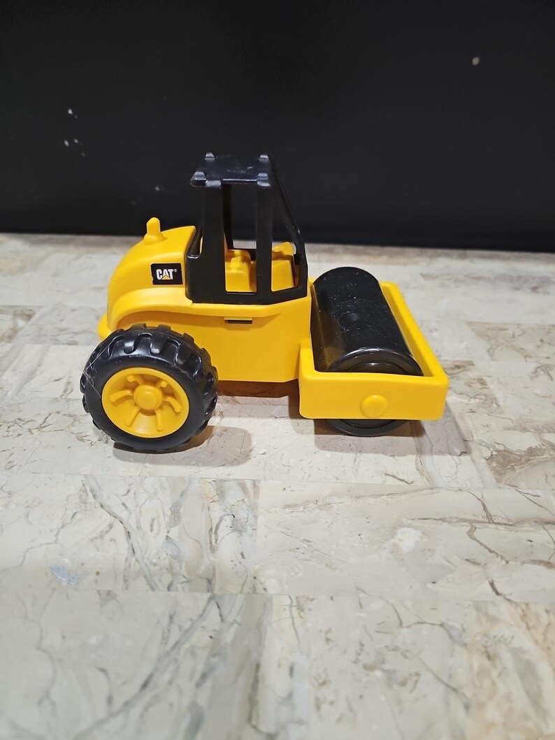 Puede incluir: Veh&iacute;culo de construcci&oacute;n de juguete amarillo con una cabina negra y un rodillo negro. El veh&iacute;culo tiene la palabra "CAT" impresa en el lateral.