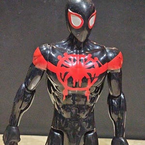 May include: A black action figure of a superhero with red accents. The figure has a black mask with white eye shapes, red shoulder pads, and a red emblem on the chest. The figure is posed with arms at its sides.
