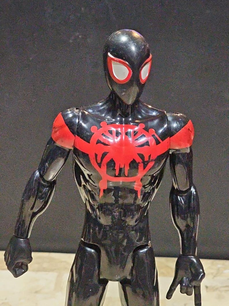 May include: A black action figure of a superhero with red accents. The figure has a black mask with white eye shapes, red shoulder pads, and a red emblem on the chest. The figure is posed with arms at its sides.