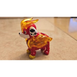 Op de afbeelding: Een rode en gele speelgoedhondfiguur met een wit gezicht, zwarte vlekken en vlamachtige accenten. De speelgoedhond staat op vier poten en heeft gele accenten op zijn poten en hoofd.