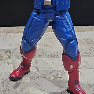 May include: A red, white, and blue action figure with blue pants and red boots. The figure is wearing a belt with a silver buckle.