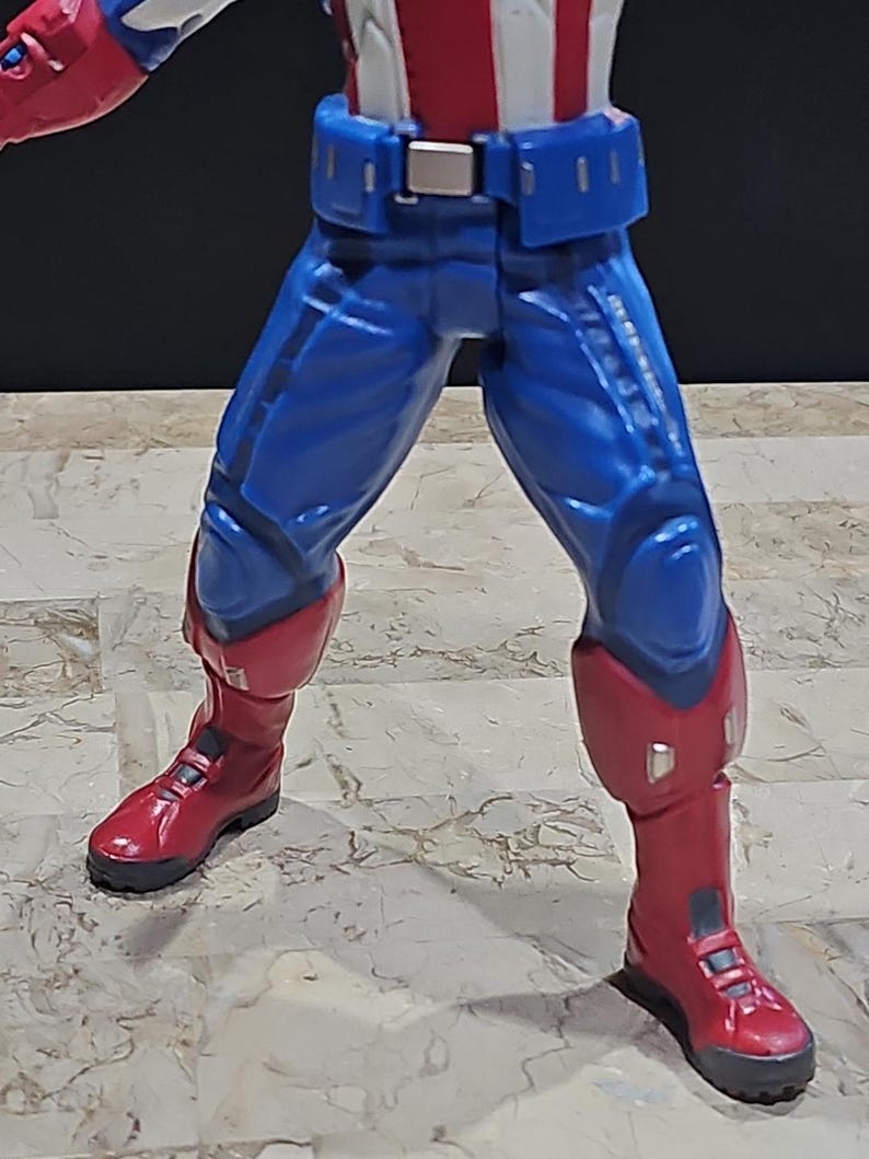 May include: A red, white, and blue action figure with blue pants and red boots. The figure is wearing a belt with a silver buckle.