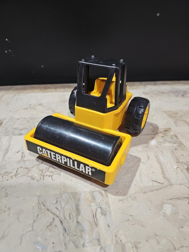 Puede incluir: Veh&iacute;culo de construcci&oacute;n de juguete amarillo con un rodillo negro y la palabra "Caterpillar" impresa en el lateral.