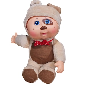 May include: A plush doll dressed in a dog costume. The doll has blue eyes, rosy cheeks, and a brown patch over one eye. It wears a red bow tie with white polka dots. The doll is sitting and giving a thumbs up.