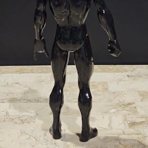 May include: A black action figure with red shoulder accents. The figure is posed with one arm extended, pointing downward. The figure's muscular physique is visible, and it stands on a patterned surface.