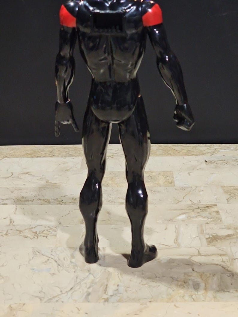 May include: A black action figure with red shoulder accents. The figure is posed with one arm extended, pointing downward. The figure's muscular physique is visible, and it stands on a patterned surface.