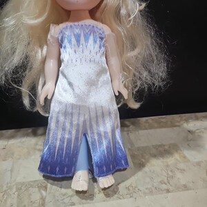 May include: A small doll with long blonde hair wearing a white and blue dress with a snowflake pattern. The doll is barefoot and has a light blue dress with a slit in the front.