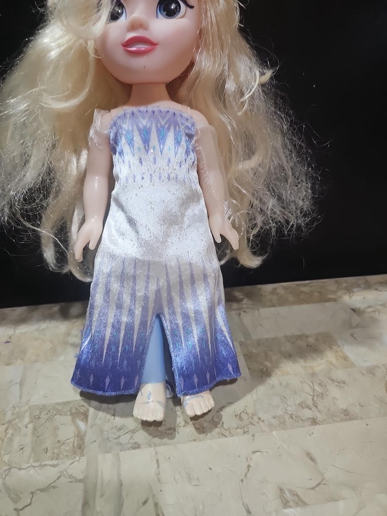 May include: A small doll with long blonde hair wearing a white and blue dress with a snowflake pattern. The doll is barefoot and has a light blue dress with a slit in the front.