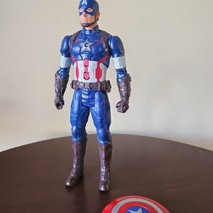 以下が含まれることがあります： 青と赤のキャプテンアメリカのフィギュアで、茶色のベルトと茶色のブーツを履いています。フィギュアは右手に盾を持っています。