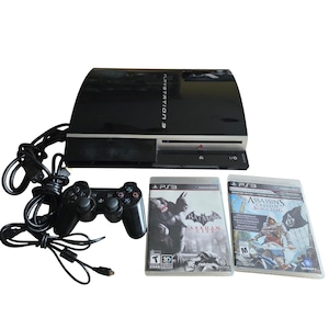 Puede incluir: Una consola de videojuegos PlayStation 3 negra con un mando inalámbrico negro a juego, un cable de alimentación y dos cajas de videojuegos tituladas Batman Arkham City y Assassin's Creed IV Black Flag, sobre un fondo blanco liso.