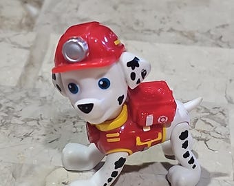 May include: A white and black dalmatian dog toy wearing a red firefighter helmet with a headlamp and a red backpack with a yellow fire emblem. The toy is made of plastic and has a cartoonish style.