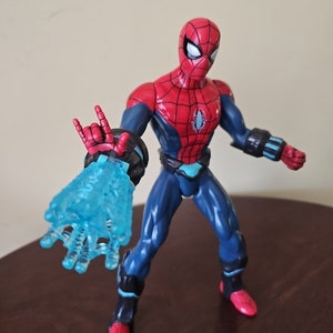 May include: A red and blue action figure of Spider-Man with a blue web-like blast effect coming from his right hand. The figure has a black spider symbol on its chest.