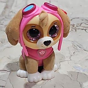 May include: A small plush toy dog wearing a pink helmet with goggles and a pink vest. The dog has brown fur, big brown eyes with pink glitter, and a pink nose. The dog is sitting on a white surface.