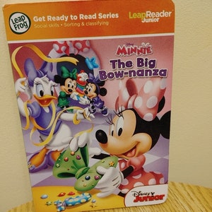 Puede incluir: Una portada de libro infantil con Minnie Mouse y Daisy Duck. El libro se titula "The Big Bow-nanza" y forma parte de la "Get Ready to Read Series" de LeapFrog. La portada está ilustrada con Minnie Mouse vistiendo un vestido rosa de lunares y un gran lazo rosa, y Daisy Duck vistiendo un vestido morado con una cinta amarilla.