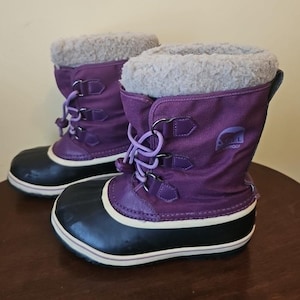 May include: A pair of purple winter boots featuring a black rubber lower shell, a white midsole, and a light gray faux-fur lining at the collar. The boots have a lace-up front with metal eyelets, purple laces, and a Sorel logo on the side.