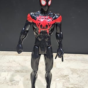 May include: A black and red action figure of a superhero. The figure has a black suit with red accents on the shoulders and chest. The mask has white eye lenses. The figure is standing on a patterned surface.