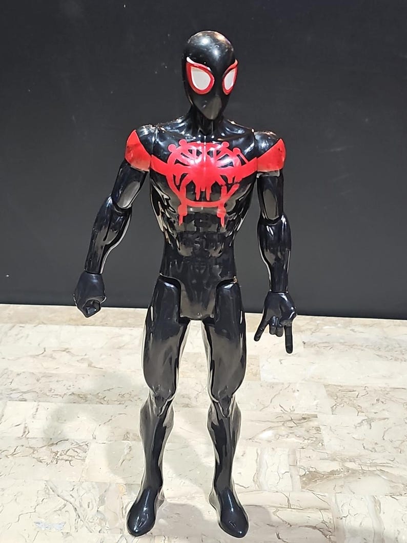 May include: A black and red action figure of a superhero. The figure has a black suit with red accents on the shoulders and chest. The mask has white eye lenses. The figure is standing on a patterned surface.