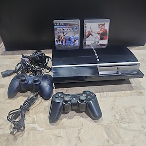 Op de afbeelding: Een zwarte PlayStation 3-console met twee zwarte controllers en twee gamecases. De gamecases zijn gelabeld met "Sports Champions" en "NCAA Football". De controllers en console staan op een lichtgekleurd oppervlak.