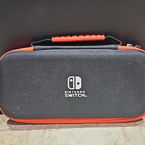 May include: Black Nintendo Switch carrying case with a red zipper and handle. The case has the Nintendo Switch logo and text on the front.
