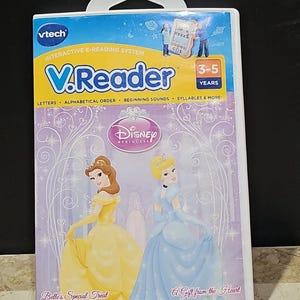 May include: VTech V.Reader interactive e-reading system for ages 3-5. The cover features Disney Princesses Belle and Cinderella, with text that reads "Belle's Special Treat" and "A Gift from the Heart". The packaging is white with a clear handle.