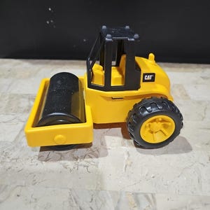 Puede incluir: Veh&iacute;culo de construcci&oacute;n de juguete amarillo con una cabina negra y un rodillo negro. El veh&iacute;culo tiene la palabra "CAT" impresa en el lateral.