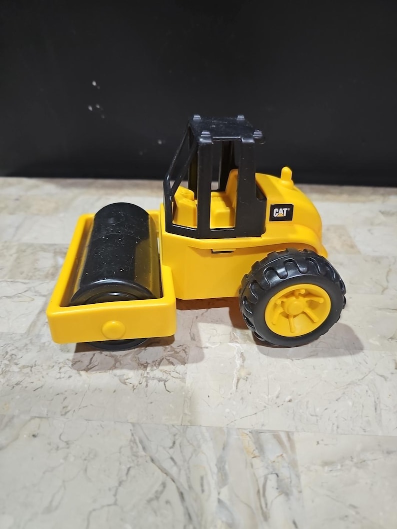 Puede incluir: Veh&iacute;culo de construcci&oacute;n de juguete amarillo con una cabina negra y un rodillo negro. El veh&iacute;culo tiene la palabra "CAT" impresa en el lateral.