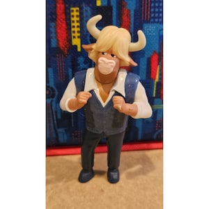 May include: A figurine of a bull-like character with a tan face, blonde hair, and white horns. The figure wears a white shirt, a blue vest, and black trousers. The figure is standing on a brown surface.