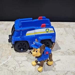 Op de afbeelding: Een blauwe en gele speelgoedpolitieauto met een rood lampje bovenop. Een bruine en gele speelgoedhond in een blauw politiekostuum staat voor de auto. De auto heeft het nummer 02 aan de zijkant.