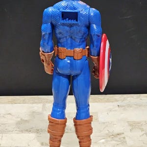 May include: A blue and red plastic action figure of Captain America, wearing a blue and red costume with a red, white, and blue shield. The figure is standing with its back to the camera, and its arms are at its sides.